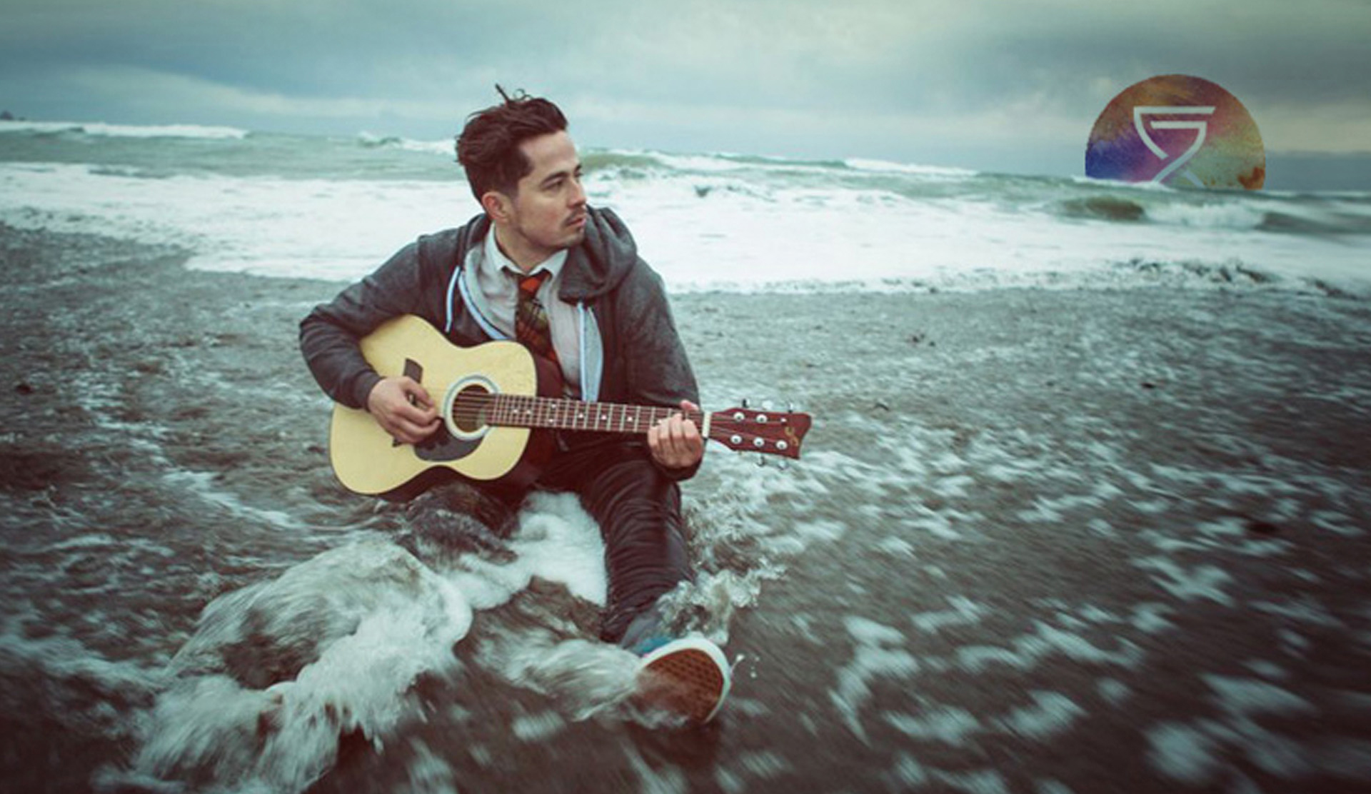 Independent artist Scott Zuniga sits in water on a rainy, windy beach. The independent singer songwriter plays the guitar because he is a guitar songwriter. The waves behind him are ominous and approaching quickly. Scott plays undisturbed.